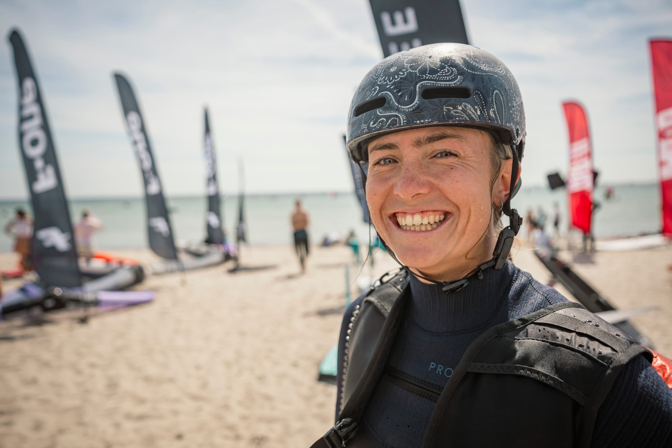 Lachende Surferin mit Helm und Neoprenanzug am Strand, im Hintergrund Fahnen der Aussteller:innen